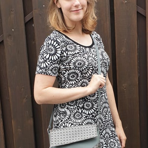 Puede incluir: Una mujer con un vestido floral en blanco y negro y un bolso bandolera gris. El bolso tiene un patr&oacute;n geom&eacute;trico en la solapa y un cuerpo gris liso. El fondo es una valla de madera marr&oacute;n oscuro.