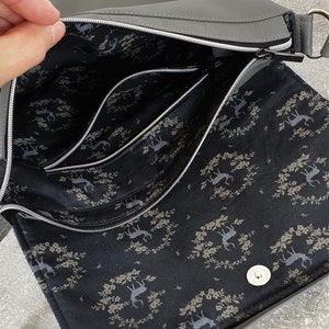 Puede incluir: Bolso bandolera gris con cierre de solapa y herrajes plateados. El interior presenta m&uacute;ltiples compartimentos con un forro de tela negra adornado con un estampado de ciervos y flores. El bolso tiene cierre de cremallera y bandolera.