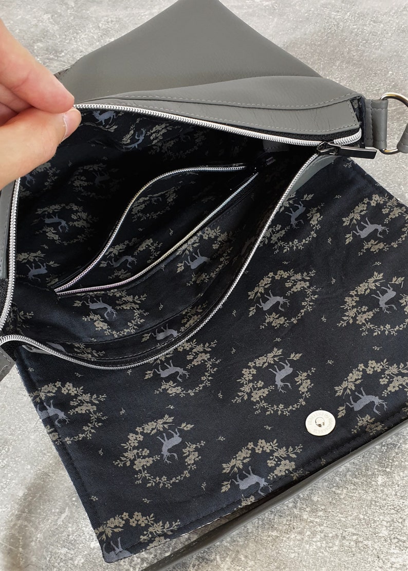 Puede incluir: Bolso bandolera gris con cierre de solapa y herrajes plateados. El interior presenta m&uacute;ltiples compartimentos con un forro de tela negra adornado con un estampado de ciervos y flores. El bolso tiene cierre de cremallera y bandolera.