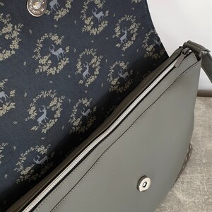 Puede incluir: Bolso bandolera gris con cremallera plateada y cierre magn&eacute;tico. El forro interior presenta una tela azul oscuro con un estampado de ciervos y coronas florales. El bolso tiene una correa gris.