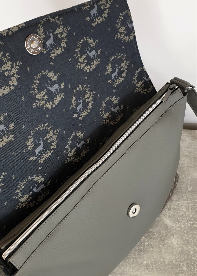 Puede incluir: Bolso bandolera gris con cremallera plateada y cierre magn&eacute;tico. El forro interior presenta una tela azul oscuro con un estampado de ciervos y coronas florales. El bolso tiene una correa gris.