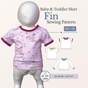 May include: A pink baby and toddler shirt with a unicorn pattern, rainbow, and heart designs. The shirt has burgundy trim on the sleeves and neckline. The image also includes the text "Baby & Toddler Shirt Fin Sewing Pattern".