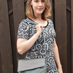 Puede incluir: Una mujer con un vestido floral en blanco y negro lleva un bolso de hombro gris. El bolso tiene un patr&oacute;n geom&eacute;trico en la solapa y un cuerpo gris liso. El fondo es una valla de madera marr&oacute;n oscuro.