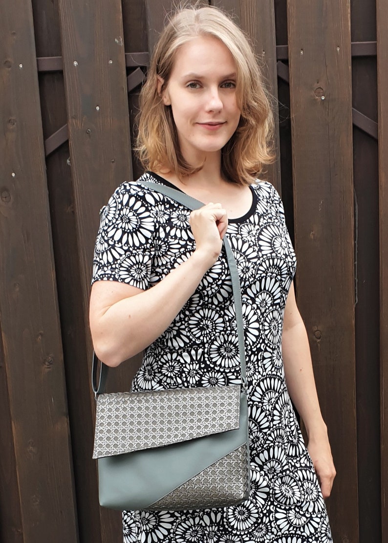 Puede incluir: Una mujer con un vestido floral en blanco y negro lleva un bolso de hombro gris. El bolso tiene un patr&oacute;n geom&eacute;trico en la solapa y un cuerpo gris liso. El fondo es una valla de madera marr&oacute;n oscuro.