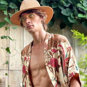 May include: A straw fedora hat and yellow-tinted sunglasses. A cream-coloured kimono with a floral pattern in red, brown, and teal. The kimono is open, revealing the torso. The background features green foliage and a wooden wall.