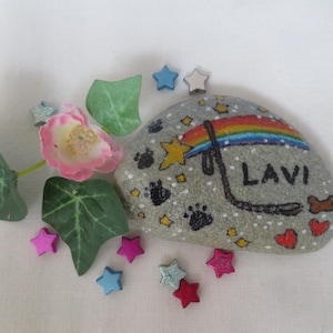 Könnte beinhalten: Ein bemalter Stein mit dem Namen "LAVI" und einem Regenbogen-Design. Der Stein ist mit Pfotenabdrücken, Sternen und Herzen verziert. Dekorative Blätter, eine rosa Blume und sternförmige Verzierungen umgeben den Stein.