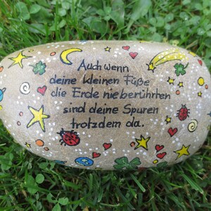 May include: A smooth, light brown river stone painted with black German text that reads: Auch wenn deine kleinen F&uuml;&szlig;e die Erde nie ber&uuml;hrten sind deine Spuren trotzdem da. The stone features colorful hand-painted stars, hearts, ladybugs, and planets.