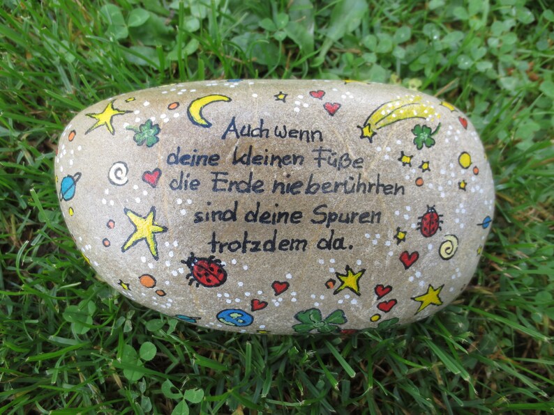 May include: A smooth, light brown river stone painted with black German text that reads: Auch wenn deine kleinen F&uuml;&szlig;e die Erde nie ber&uuml;hrten sind deine Spuren trotzdem da. The stone features colorful hand-painted stars, hearts, ladybugs, and planets.