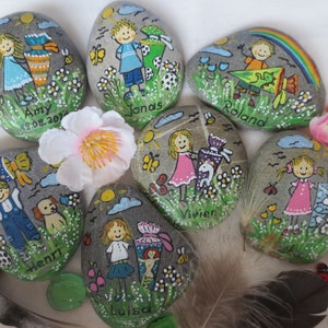 May include: Hand-painted decorative stones featuring children, flowers, butterflies, and rainbows. Each stone has a child's name and a date. The artwork includes a variety of colors and whimsical designs, perfect for gifts or decor.