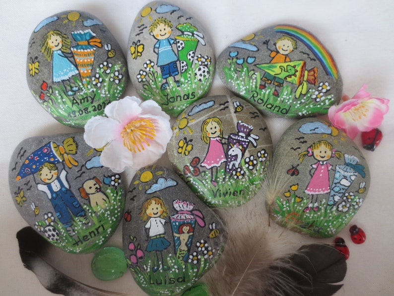May include: Hand-painted decorative stones featuring children, flowers, butterflies, and rainbows. Each stone has a child's name and a date. The artwork includes a variety of colors and whimsical designs, perfect for gifts or decor.