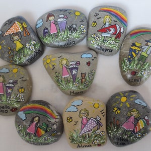 May include: Hand-painted decorative rocks featuring children with school cones, flowers, and rainbows. Each rock has a unique design with names like "Mischa" and "Isabella 2020". The rocks are painted with bright colors and whimsical details.