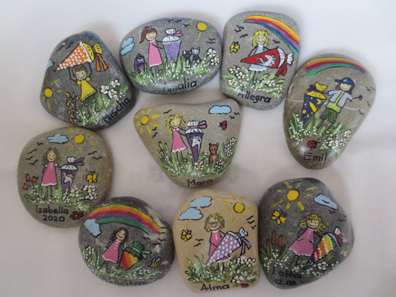 May include: Hand-painted decorative rocks featuring children with school cones, flowers, and rainbows. Each rock has a unique design with names like "Mischa" and "Isabella 2020". The rocks are painted with bright colors and whimsical details.