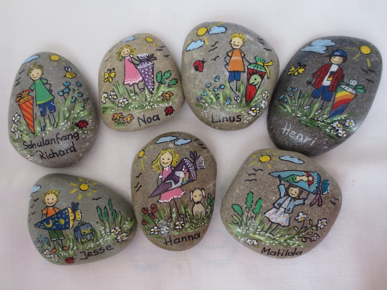 May include: Hand-painted decorative stones featuring children with backpacks and the names Richard, Noa, Linus, Henri, Jesse, Hanna, and Matilda. Each stone has a unique design with flowers, suns, and insects.