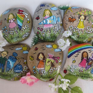 May include: Hand-painted decorative stones featuring children, flowers, and insects. Each stone has a child's name, a school cone, and a whimsical design. The stones are painted with bright colors and are set against a background of green grass and white flowers.
