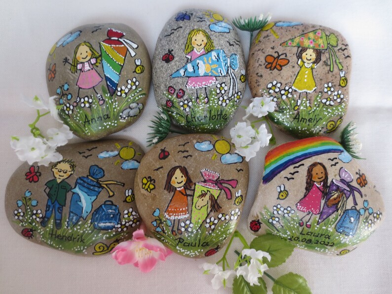 May include: Hand-painted decorative stones featuring children, flowers, and insects. Each stone has a child's name, a school cone, and a whimsical design. The stones are painted with bright colors and are set against a background of green grass and white flowers.
