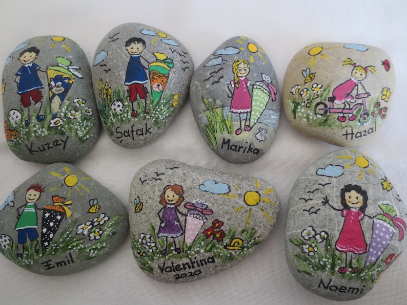 May include: Hand-painted decorative stones featuring children with school cones, flowers, and sunny skies. Each stone has a child's name, such as Kuzey, Safak, and Marika. The artwork includes details like bees, birds, and a bicycle.
