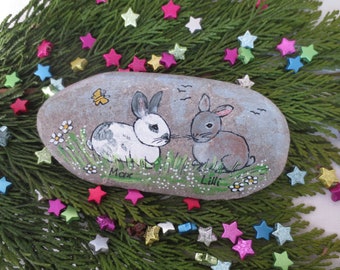 May include: Hand-painted stone with two rabbits, one white with black spots and one brown, with the names Max and Lilli written below them. The stone is surrounded by green grass and white flowers.
