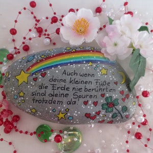 May include: A smooth gray river stone painted with a colorful rainbow, yellow stars, a crescent moon, red hearts, and a green four-leaf clover. The German text reads: Auch wenn deine kleinen F&uuml;&szlig;e die Erde nie ber&uuml;hrten sind deine Spuren trotzdem da.