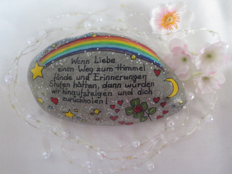 May include: A smooth gray river stone painted with a colorful rainbow, yellow stars, a yellow crescent moon, red hearts, and a green four-leaf clover. The German text reads: Wenn Liebe einen Weg zum Himmel f&auml;nde und Erinnerungen Stufen h&auml;tten, dann w&uuml;rden wir hinaufsteigen und dich zur&uuml;ckholen!