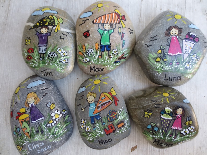 May include: Hand-painted decorative stones featuring children with school-themed illustrations. Each stone has a unique design, including children with school bags, flowers, and the names Tim, Max, Luna, Elisa, Nico, and Mona. The stones are gray with colorful artwork.