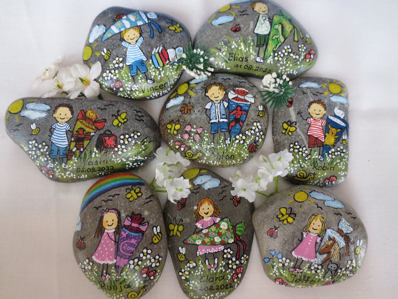 May include: Hand-painted decorative rocks featuring children with school-themed illustrations. Each rock has a unique design with names and dates, including Vincent, Elias, Yasin, Anton, Robin, Ronja, Clara, and Jette. The rocks are adorned with flowers, butterflies, and other whimsical details.