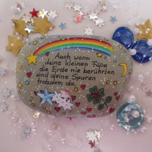 May include: A smooth gray stone painted with a colorful rainbow, a yellow crescent moon, small red hearts, and a green four-leaf clover. The German text reads: Auch wenn deine kleinen F&uuml;&szlig;e die Erde nie ber&uuml;hrten, sind deine Spuren trotzdem da.