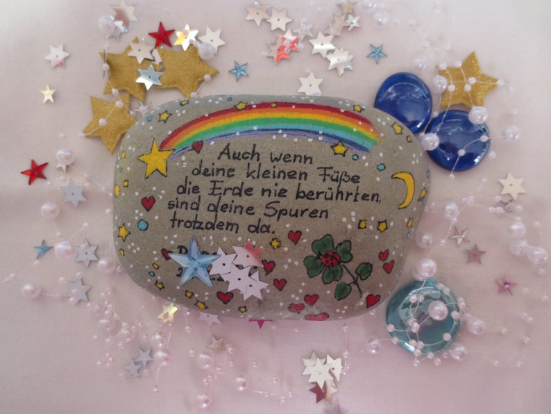May include: A smooth gray stone painted with a colorful rainbow, a yellow crescent moon, small red hearts, and a green four-leaf clover. The German text reads: Auch wenn deine kleinen F&uuml;&szlig;e die Erde nie ber&uuml;hrten, sind deine Spuren trotzdem da.