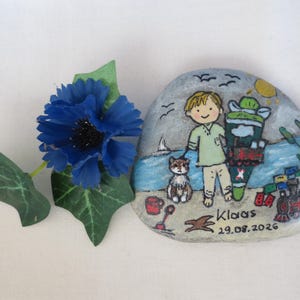 May include: A hand-painted stone featuring a beach scene with a child, a cat, and a toy train. The artwork includes the name "Klaas" and the date "19.08.2026". A blue flower with green leaves is placed on the left.