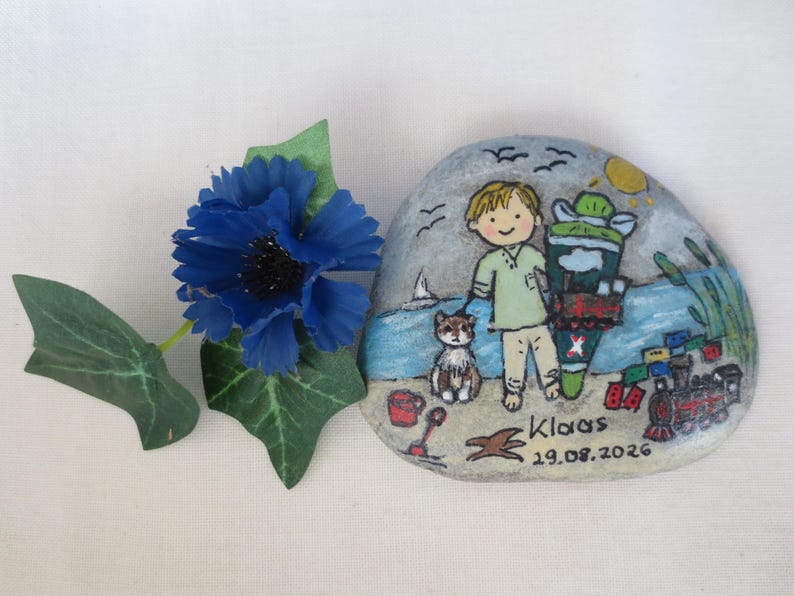 May include: A hand-painted stone featuring a beach scene with a child, a cat, and a toy train. The artwork includes the name "Klaas" and the date "19.08.2026". A blue flower with green leaves is placed on the left.