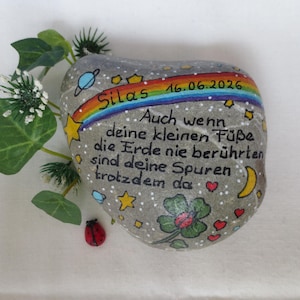 May include: A grey painted stone featuring a rainbow, stars, planets, a four-leaf clover, and a ladybug. The text reads: Silas 16.06.2026, Auch wenn deine kleinen F&uuml;&szlig;e die Erde nie ber&uuml;hrten sind deine Spuren trotzdem da. Includes green leaves.