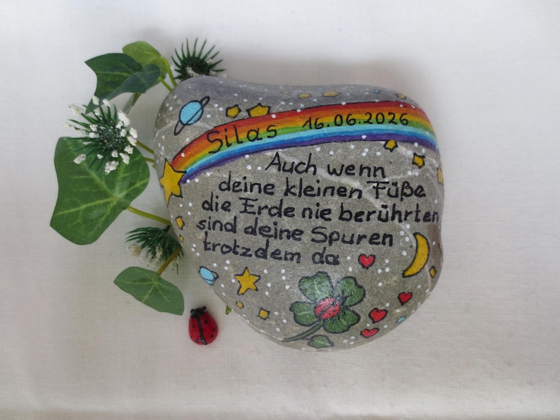 May include: A grey painted stone featuring a rainbow, stars, planets, a four-leaf clover, and a ladybug. The text reads: Silas 16.06.2026, Auch wenn deine kleinen F&uuml;&szlig;e die Erde nie ber&uuml;hrten sind deine Spuren trotzdem da. Includes green leaves.