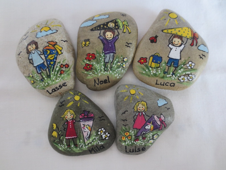 May include: Five hand-painted stones with illustrations of children, school cones, and flowers. Each stone features a child's name: Lasse, Noel, Luca, Mila, and Luise. The artwork includes a sun, clouds, and a ladybug.