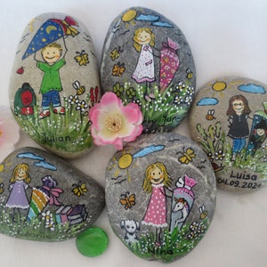 May include: Hand-painted decorative rocks featuring children, flowers, butterflies, and the sun. Each rock has a unique design, including names like "Julian" and "Luisa", and a school-themed motif. The rocks are gray with colorful illustrations.