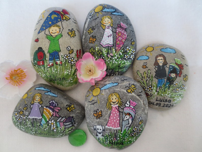 May include: Hand-painted decorative rocks featuring children, flowers, butterflies, and the sun. Each rock has a unique design, including names like "Julian" and "Luisa", and a school-themed motif. The rocks are gray with colorful illustrations.