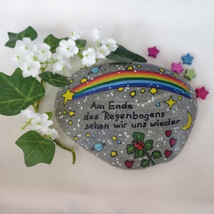 May include: A gray painted stone featuring a colorful rainbow, yellow stars, a crescent moon, planets, a ladybug on a green clover, and red hearts. The text reads: Am Ende des Regenbogens sehen wir uns wieder. White flowers are positioned to the left.