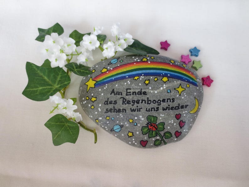 May include: A gray painted stone featuring a colorful rainbow, yellow stars, a crescent moon, planets, a ladybug on a green clover, and red hearts. The text reads: Am Ende des Regenbogens sehen wir uns wieder. White flowers are positioned to the left.