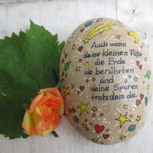 May include: A smooth, oval-shaped tan stone painted with black German text that reads, "Auch wenn deine kleinen F&uuml;&szlig;e die Erde nie ber&uuml;hrten sind deine Spuren trotzdem da." The stone features colorful hand-painted stars, hearts, and planets.