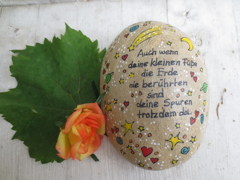 May include: A smooth, oval-shaped tan stone painted with black German text that reads, "Auch wenn deine kleinen F&uuml;&szlig;e die Erde nie ber&uuml;hrten sind deine Spuren trotzdem da." The stone features colorful hand-painted stars, hearts, and planets.
