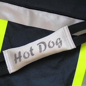 Könnte beinhalten: Ein weißes Canvas-Hundespielzeug mit schwarzen Gurtbändern und dem aufgedruckten Text "Hot Dog" auf der Vorderseite. Das Spielzeug liegt auf einem schwarzen und gelben Stoffhintergrund.