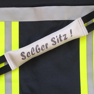 Könnte beinhalten: Ein weißes Hundetrainingstool mit schwarzen Riemen und dem aufgedruckten Text "Sitz!" Das Tool liegt auf einer schwarzen Oberfläche mit gelben und silbernen reflektierenden Streifen.