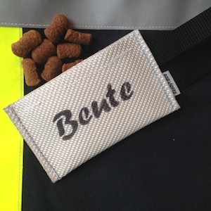 Könnte beinhalten: Ein weißes Feuerwehrschlauch-Etikett mit dem Namen "Bente" in schwarzer, kursiv geschriebener Schrift. Das Etikett ist an einem schwarzen Band befestigt.