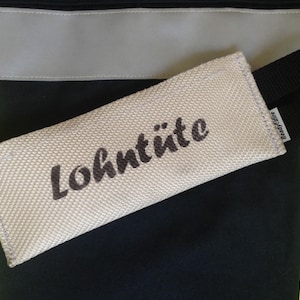 Könnte beinhalten: Ein beiger Feuerwehrschlauch mit dem aufgedruckten Wort "Lohntüte". Der Schlauch hat einen schwarzen Riemen mit dem aufgedruckten Wort "Readyfire".