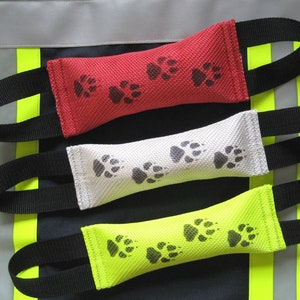 Könnte beinhalten: Drei Hundespielzeuge in Rot, Weiß und Gelb mit schwarzen Pfotenabdrücken. Jedes Spielzeug hat einen schwarzen Riemengriff.