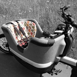 以下が含まれることがあります： グレーのカーゴバイクで、模様のあるシートと側面に「Gazelle」という文字が印刷されています。