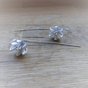 Op de afbeelding: Een paar zilveren oorhangers met een delicaat bloemenmotief. De bloemen hebben een witte afwerking en gouden accenten.