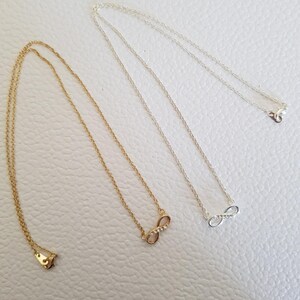 Könnte beinhalten: Zwei Halsketten mit einem Unendlichkeitszeichen, eine in Gold und eine in Silber. Beide Halsketten haben ein kleines Unendlichkeitszeichen mit einer Reihe kleiner, klarer Steine.