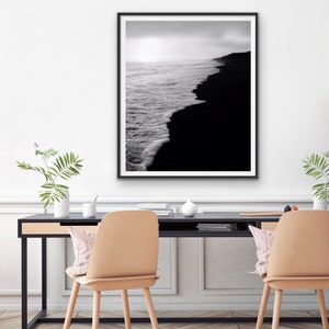May include: Black and white photograph of a black sand beach with waves crashing on the shore. The photo is framed in a black frame and hanging on a white wall.