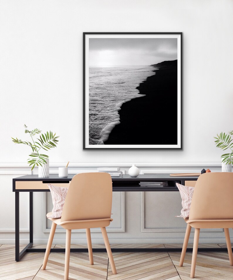 May include: Black and white photograph of a black sand beach with waves crashing on the shore. The photo is framed in a black frame and hanging on a white wall.