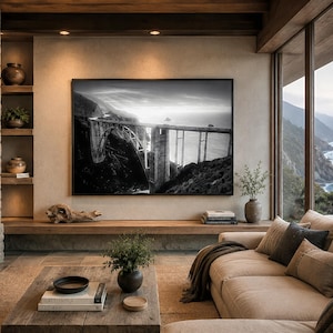 May include: A large rectangular black and white wall art print depicting a bridge over a coastal cliffside, mounted on a beige wall above a wooden shelf in a living room space featuring a stone fireplace and a beige sectional sofa.
