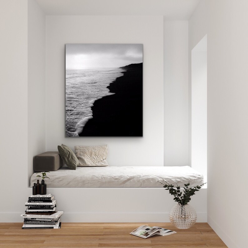 May include: Black and white photograph of a beach with waves crashing on the shore. The sand is black and the water is white. The photo is taken from a low angle, looking up at the sky.